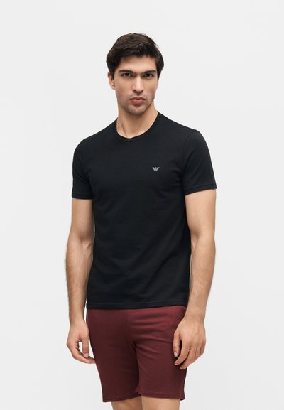 Άνδρας που φοράει μαύρο κοντομάνικο μπλουζάκι με μικρό λογότυπο στο στήθος και μπορντό σορτς, στέκεται μπροστά σε απλό ανοιχτό φόντο.