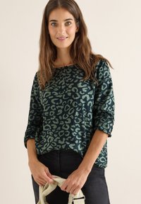 Pullover con stampa leopardo verde, con maniche lunghe, colletto rotondo e polsini arrotolati, abbinato a pantaloni scuri, mentre tiene una leggera tracolla.