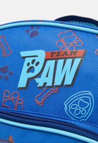 Mėlynas audinio kuprinė, išskirtinai papuošta "TEAM PAW" logotipu, letenų atspaudais ir kauliukų grafikomis raudonos ir violetinės spalvų. Yra užtrauktukas ir kontrastinga apdaila.