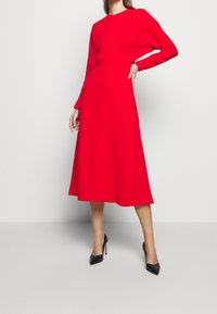 Vestido rojo de manga larga con un torso ajustado y falda midi acampanada, hecho de un tejido suave. Combinado con tacones negros de punta afilada.