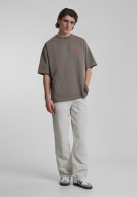 Camiseta gris de gran tamaño con un pequeño logo naranja, combinada con pantalones blancos holgados y zapatillas grises con acentos negros.