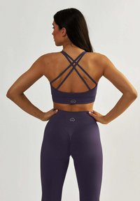 Fioletowy stanik sportowy z krzyżującymi się ramiączkami i detalem z logo, w połączeniu z wysokimi fioletowymi legginsami z marszczonym akcentem z tyłu.