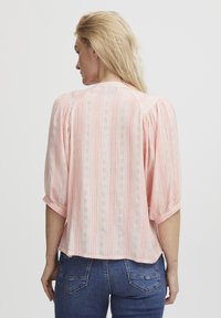 Blusa rosa chiaro con righe testurizzate, maniche a sbuffo e scollatura rotonda. Indossata con jeans blu, che mostrano una vestibilità comoda.