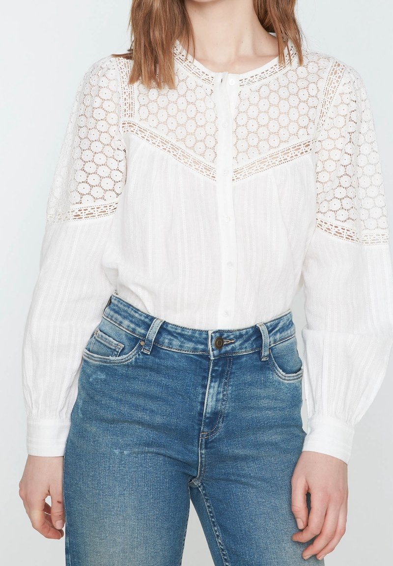 Biała bluzka z koronkowymi zdobieniami na ramionach, zapinana na guziki z przodu, z długimi rękawami. Połączona z wysokim stanem niebieskimi jeansami.
