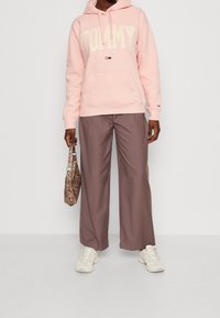 Sudadera rosa claro con el texto "TOMMY JEANS", bolsillo frontal, combinada con pantalones de pierna ancha marrones y un bolso estampado. Zapatillas blancas completan el look.