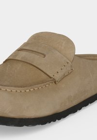 Ανοιχτόκαφέ σουέντ παπούτσι τύπου slip-on με μαύρη λαστιχένια σόλα και λεπτομέρεια με λουράκι τύπου penny στο πάνω μέρος, εμφανιζόμενο υπό ελαφριά γωνία.
