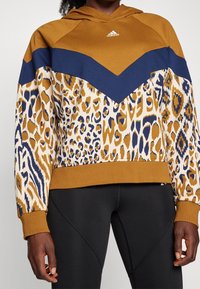 Mikina s kapucí s horní částí v hnědé barvě, tmavě modrým vzorem ve tvaru šipky a spodní částí s leopardím potiskem v žluté a modré barvě.