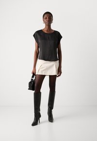 Schwarzes Kurzarmoberteil, cremefarbenen Minirock mit Falten, schwarze knielange Lederstiefel mit spitzem Zeh und Reißverschlussdetails, hält eine schwarze Tasche.