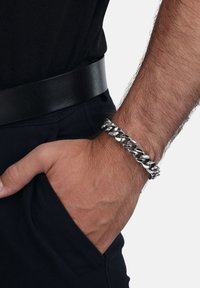 Pulsera de cadena de plata con eslabones entrelazados, acabado pulido, llevada en una muñeca. Cinturón negro mate visible en la cintura. Atuendo casual.