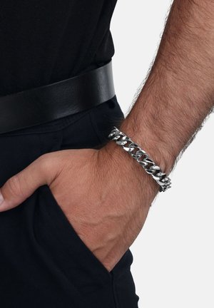 Silver chain bracelet with interlocking links, polished finish, worn on a wrist. Matte black belt visible at the waist. Casual attire.