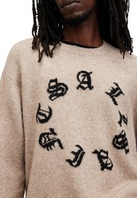 Beiger Strickpullover mit einem kreisförmigen Muster aus schwarzen, stilisierten Buchstaben. Strukturiertes Material mit Rundhalsausschnitt und lässiger Passform.