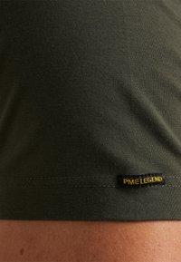 Pogled izbliza na tamnozelenu tkaninu s prošivenim rubom i malom crnom etiketom na kojoj piše "PME LEGEND" žutim slovima.
