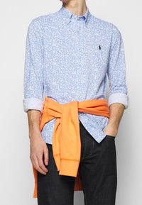 Jasnoniebieska koszula w kwiaty na guziki z białym wzorem, podwinięte rękawy. Jasnopomarańczowa bluza z kapturem zawiązana wokół talii, ciemne dżinsy.