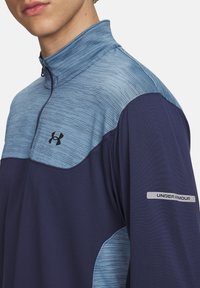 Tumesinine ja helesinine veerandzipperiga pullover, millel on tekstuuriga kangas, must logo rinnal ja hõbedane aktsent varrukal.