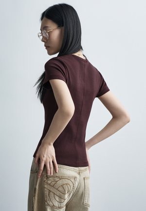 Femme aux longs cheveux noirs portant des lunettes, un haut ajusté bordeaux à manches courtes et un pantalon beige avec des poches arrière brodées, posant de profil.