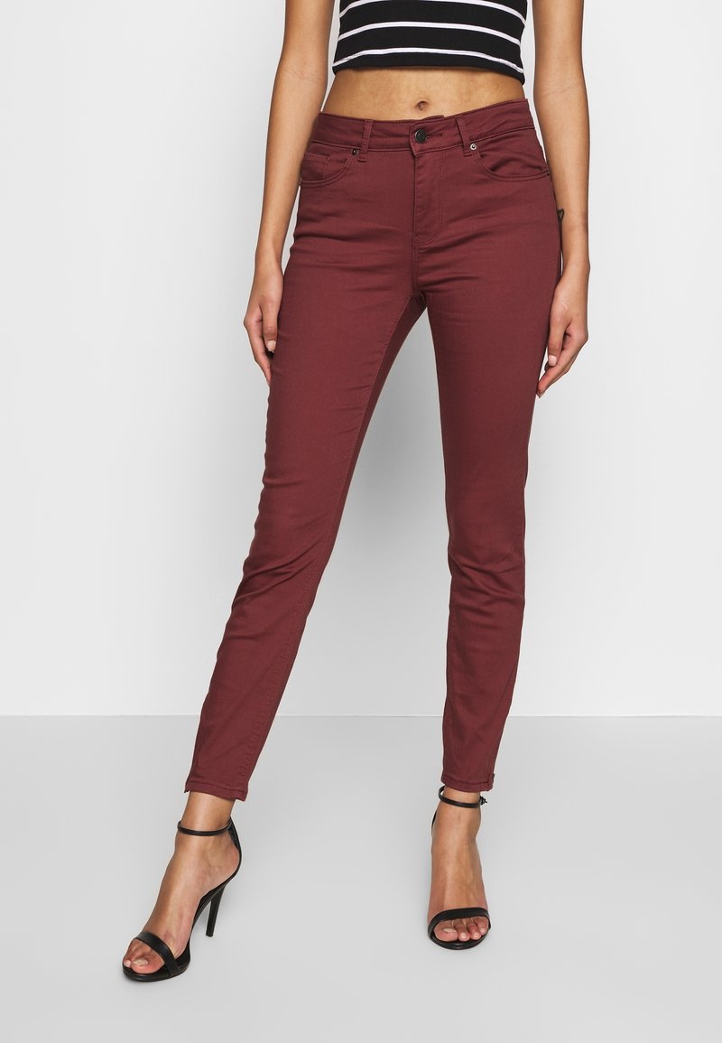 Jeans skinnies burdeos con una textura suave, cinco bolsillos y cierre de botón en la parte delantera, combinados con tacones altos negros con tiras.