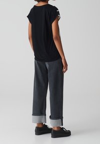 Osoba w czarnym, przerośniętym t-shircie, ciemnych, szerokich jeansach z mankietami i czarnych sznurowanych butach, stojąca na tle jednolitego, jasnego tła.