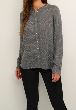 Frau trägt eine graue Bluse mit Knöpfen und weißem geometrischem Muster sowie schwarze Hose, steht vor einem schlichten hellen Hintergrund.