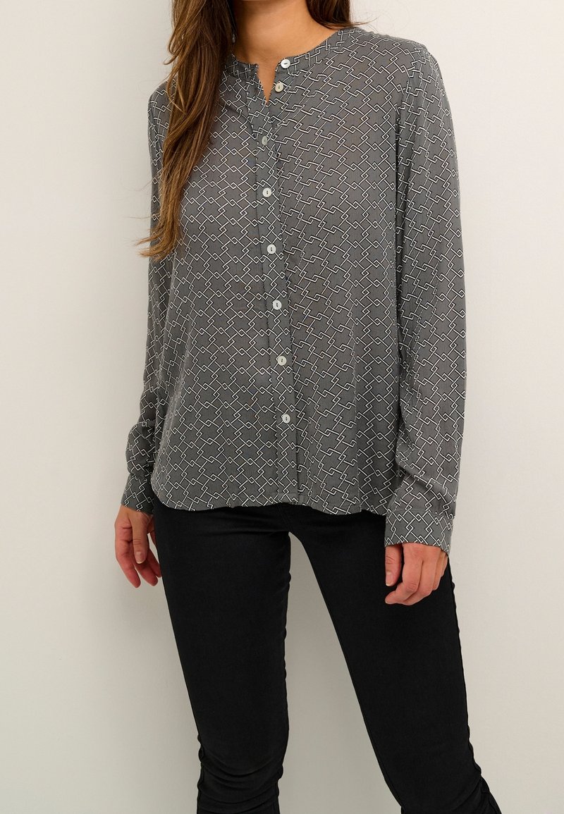 Femme portant une blouse grise à boutons avec un motif géométrique blanc et un pantalon noir, debout devant un fond clair uni.