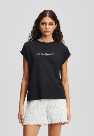Schwarzes Baumwoll-T-Shirt mit aufgerollten Ärmeln und einem weißen gestickten Design auf der Vorderseite. Kombiniert mit hellen, strukturierten Tweed-Shorts.