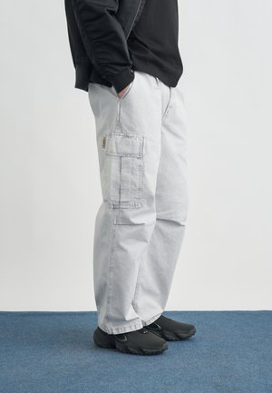 Person trägt hellgraue Cargohosen mit Seitentasche und schwarze Schuhe und steht auf blauem Teppichboden vor weißem Hintergrund.