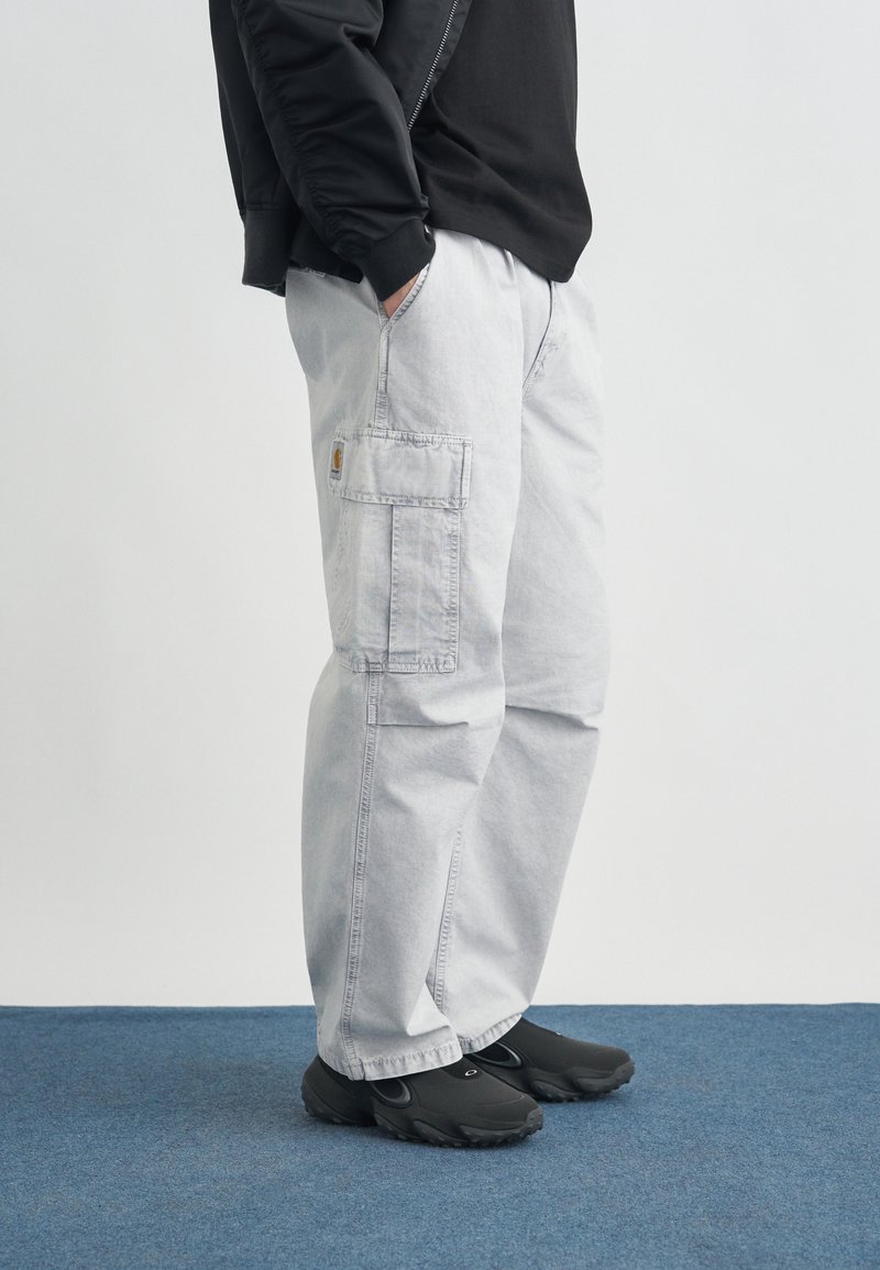 Person trägt hellgraue Cargohosen mit Seitentasche und schwarze Schuhe und steht auf blauem Teppichboden vor weißem Hintergrund.