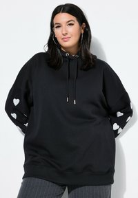 Sudadera negra hecha de tela suave, con un bolsillo canguro y patrones de corazones plateados en las mangas. Cuerda ajustable con puntas metálicas.