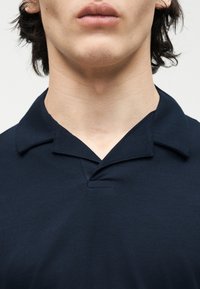 Νεαρός με ελαφρώς ανοιχτά χείλη που φοράει σκούρο μπλε πουκάμισο με γιακά, μπροστά σε απλό ανοιχτό φόντο.