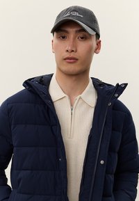 Νέος άνδρας που φοράει μπλε μπουφάν puffer πάνω από ένα κρεμ πουλόβερ με φερμουάρ και μαύρο καπέλο του μπέιζμπολ με κεντημένο το "Les Deux".