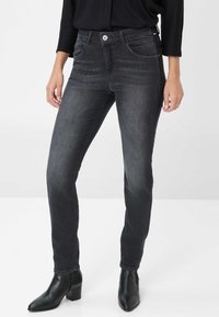 Femme portant un jean skinny gris foncé et des bottines noires, se tenant contre un fond clair et uni, les mains détendues sur les côtés.