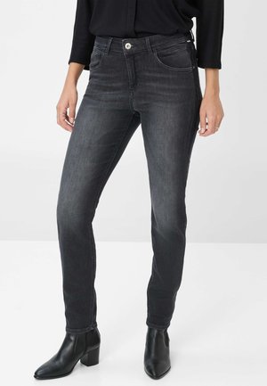 Frau in dunkelgrauen Skinny Jeans und schwarzen Stiefeletten, die vor einem schlichten, hellen Hintergrund steht, mit entspannten Händen an den Seiten.