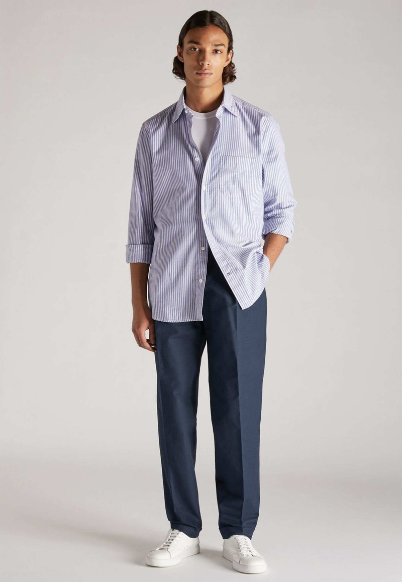 Camisa de botones a rayas en azul claro y blanco, combinada con pantalones oscuros de color navy y zapatillas blancas. La camisa tiene un bolsillo y las mangas enrolladas.