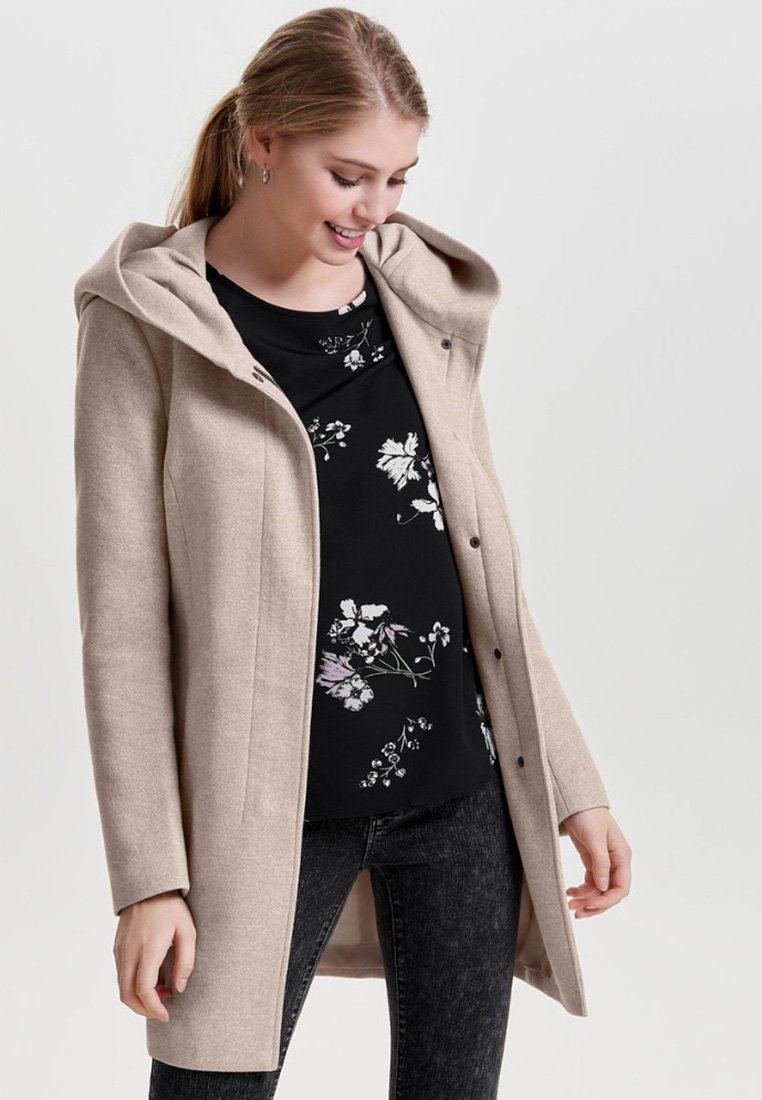 Beige hettegenser med en myk tekstur, med en knappeåpning og sidelommer, båret over en svart topp med blomsterdrømmer.