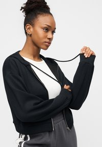 Μαύρο cropped hoodie με φερμουάρ μπροστά, φαρδιά μανίκια και ρυθμιζόμενο κορδόνι. Συνδυασμένο με λευκό τοπ και γκρι φόρμας.