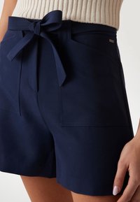 Námořnické modré šortky vyrobené z hladkého materiálu, s přední šňůrkou, dvěma velkými náprsními kapsami a strukturovaným střihem s minimem švů.