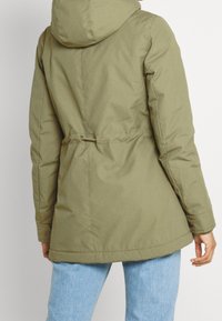 Olivgrön vadderad parkas med huva, markerad midja och ärmslut med knappar. Har en slät yta och en lätt rundad fåll.