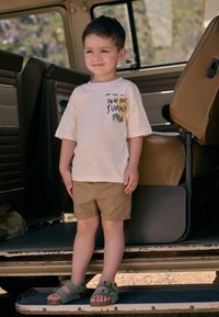 Beige T-Shirt mit kurzen Ärmeln und Grafikdruck in Kombination mit khakifarbenen Shorts und grünen Sandalen. Stehend auf einer Fahrzeugstufe, mit einem strukturierten Hintergrund.