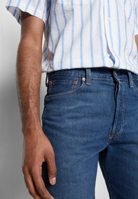 Jeans de ganga azul com corte reta, apresentando ferragens em cobre e um cós cosido. Combinados com uma camisa branca com riscas azuis.