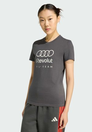 Jonge vrouw met donker haar in een knot, gekleed in een grijs Revolut F1 Team T-shirt en donkere grijze Adidas broek met rode strepen.