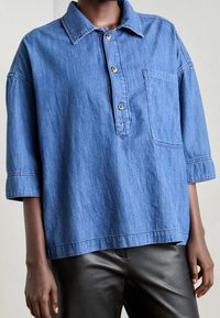 Blaues Jeanshemd mit Kragen, kurzen Ärmeln, einer Brusttasche und Knopfverschluss am Hals. Glatte Textur, entspannter Sitz.