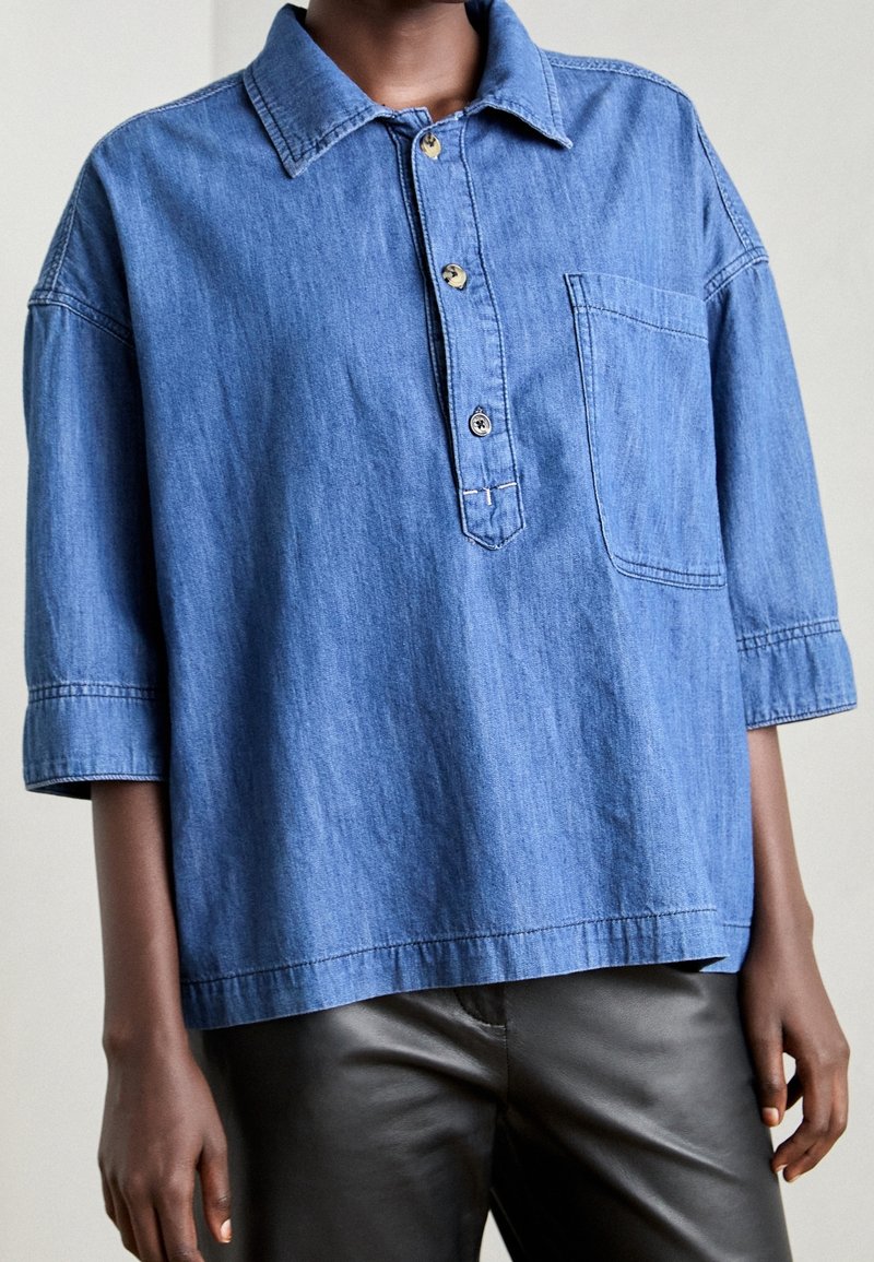 Blaues Jeanshemd mit Kragen, kurzen Ärmeln, einer Brusttasche und Knopfverschluss am Hals. Glatte Textur, entspannter Sitz.