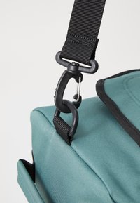 Détail d'un sac en tissu bleu-vert avec une sangle et un clip noirs. Présente une surface texturée et une poche latérale. Des éléments matériels robustes sont visibles.