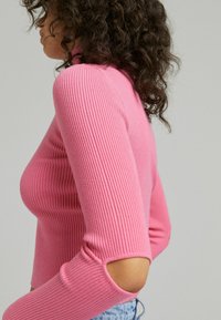 Pink ribbed knit top with a high neckline and cut-out accents on the sleeves, paired with light blue denim. Smooth texture.
