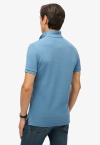 Polo shirt azzurro chiaro con un design texturizzato, colletto dritto e maniche arrotolate. Presenta fessure laterali all'orlo. Vista posteriore mostrata.