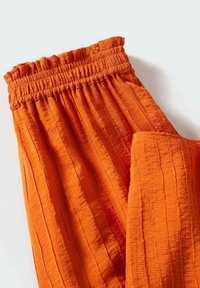 Oranžové, zmačkané látkové kalhoty s elastickým pasem, se zdeformovanými jemnými vertikálními pruhy a volným střihem. Viditelná textura.