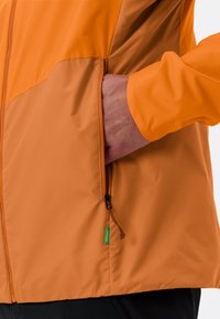 Oranje lichtgewicht jack met een soepele textuur, ritszak en verstelbare zoom. Beschikt over een groene tag aan de onderlinkerzijde voor identificatie.