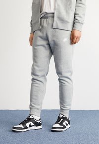 Šedé tepláky z měkké tkaniny, s elastickým pasem na šňůrku a malým bílým logem Nike na dolní části nohy. V páru s černobílými teniskami.