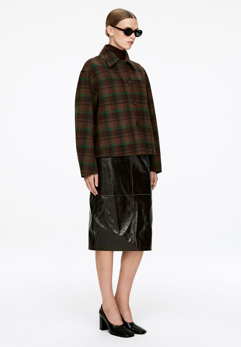 Manteau à carreaux marron et vert avec un col, jupe en cuir noir brillant et chaussures à talons carrés noires. Design ajusté.