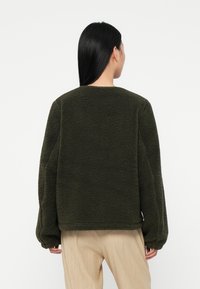 Woman with long black hair wearing a dark green fleece jacket and light beige pants, facing away against a plain white background.
