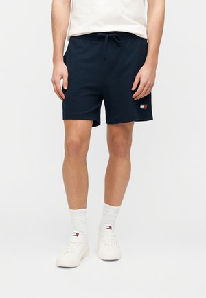 Homme portant un short bleu marine à cordon, des chaussettes blanches et des baskets blanches avec logo, debout devant un fond uni.